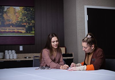 Two women sit at a table. One is instructing the other on paperwork spread out in front of them.