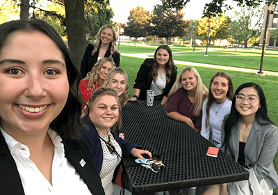 A group of nine girls pose for a selfie while sitting around a picnic table.