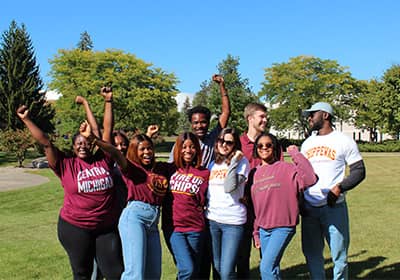 A group of communication graduate students outside, cheering with their hands up.