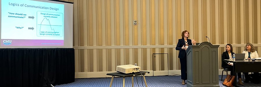 Professor Elizabeth Carlson standing at a podium speaking at a conference. You can see two other panelists seated at a table next to her. You can also see a slide from her PowerPoint displayed on a large screen behind her.