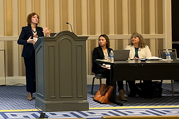 Professor Elizabeth Carlson standing at a podium speaking at a conference. You can see two other panelists seated at a table next to her.