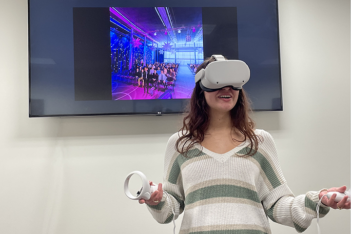 Student using virtual reality goggles to practice speaking in front of a large audience. You can see the audience that she sees displayed on a monitor behind her.