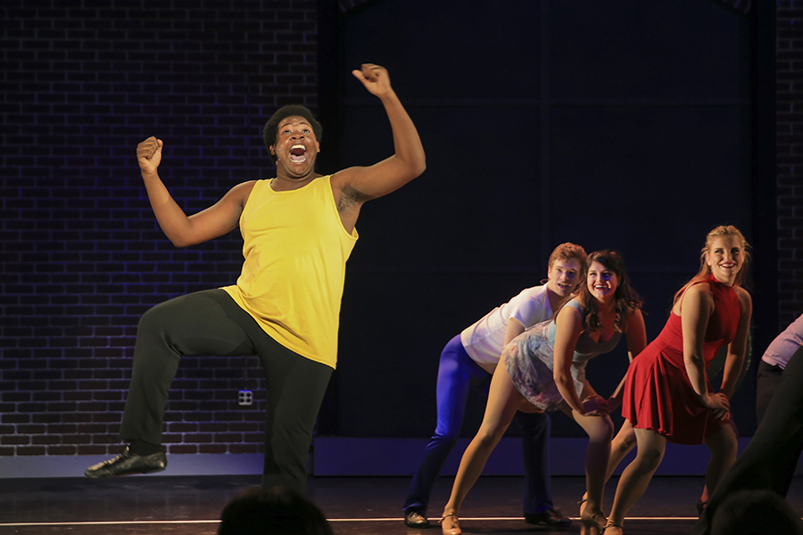 A man in a yellow tank top sings and dances with cast members behind him.