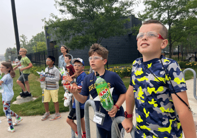 6 middle school students are outside. They are all facing towards the camera and looking up to the sky. They are holding bottle rockets.