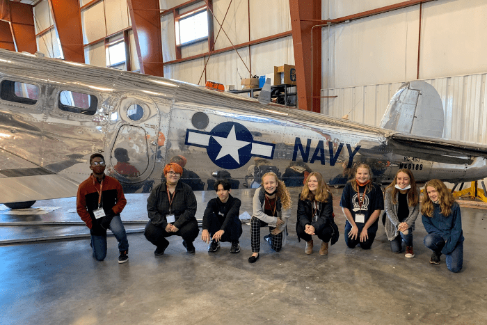 eight middle school students are kneeling in front of a WWII navy airplane.