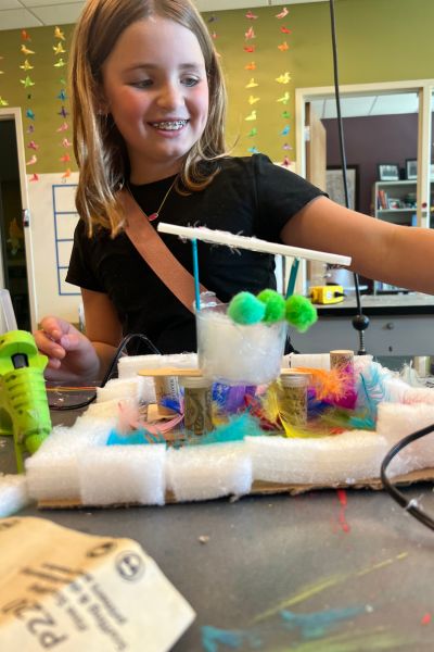 Child is smiling at something out of the frame. In front of them is a creation make of recycled materials.