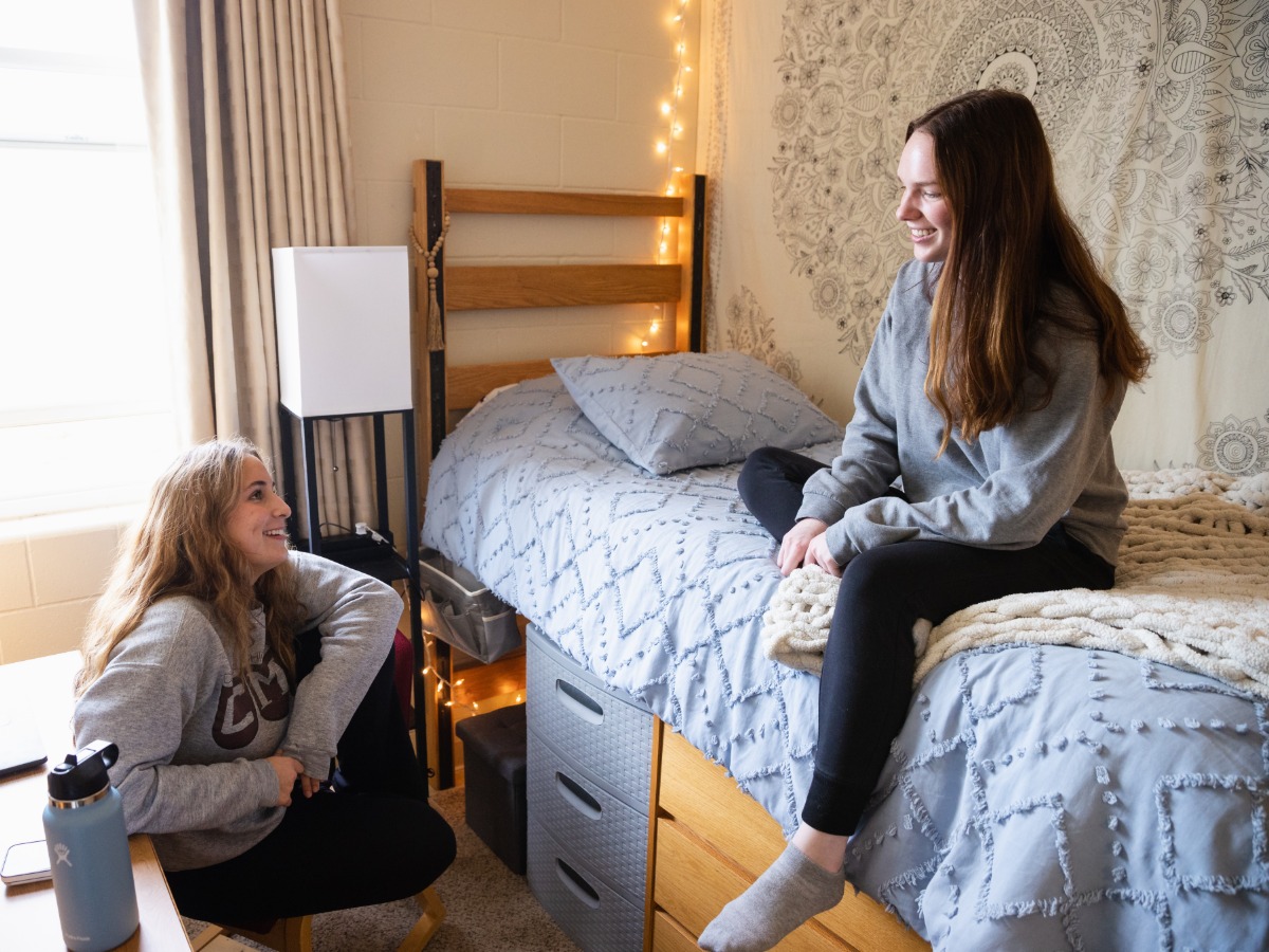 Two students sitting in a dorm room talking to each other.