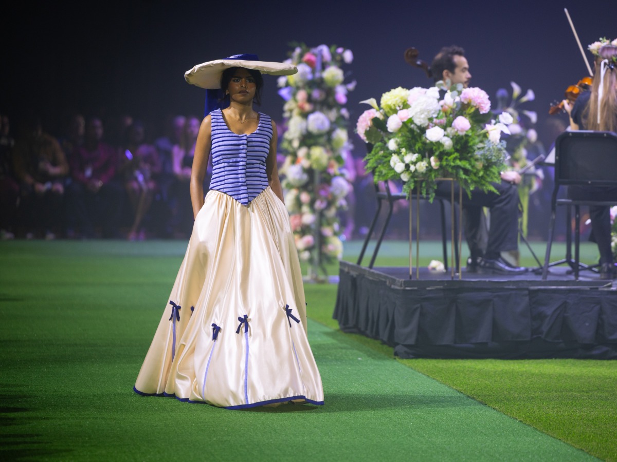 A woman in a long dress and hat walking on a green runway during Threads Fashion Show.