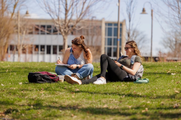 2026-06-03-Students-sitting-grass-area-01-720x480 Two female students sitting on grass with the Bovee University Center behind them.