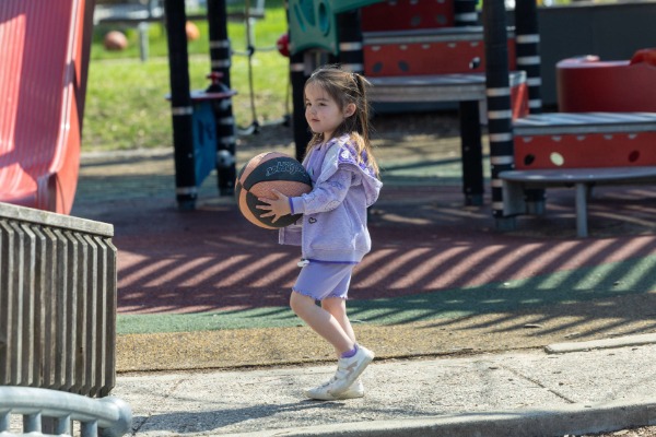 A young child outside at a playground carrying a basketball.