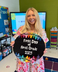 Kayley Oyster holding up a First Day of First Grade sign inside a classroom.