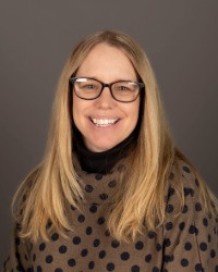 Headshot of Kristen Kostlienley wearing a polka dot shirt and glasses against a grey background.