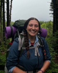Sophie Perry wearing a hiking gear backpack in front of a lake and trees.