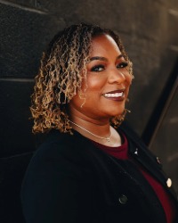 Headshot of Tanisha Bolden wearing a black cardigan against a dark grey brick wall.