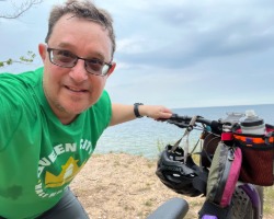 2026-Lubig-bike-01-250x200 Joe Lubig posting with his bike with sand and a lake behind him.