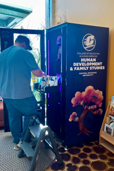 Riley Justis standing in front of the blue book vending machine, loading books inside on a tray.