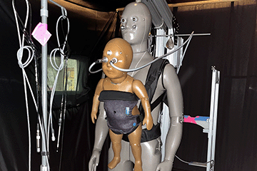 One adult sized manikin with a baby sized manikin strapped to it's chest. The manikins are inside of a cold environment chamber testing for cold climate.
