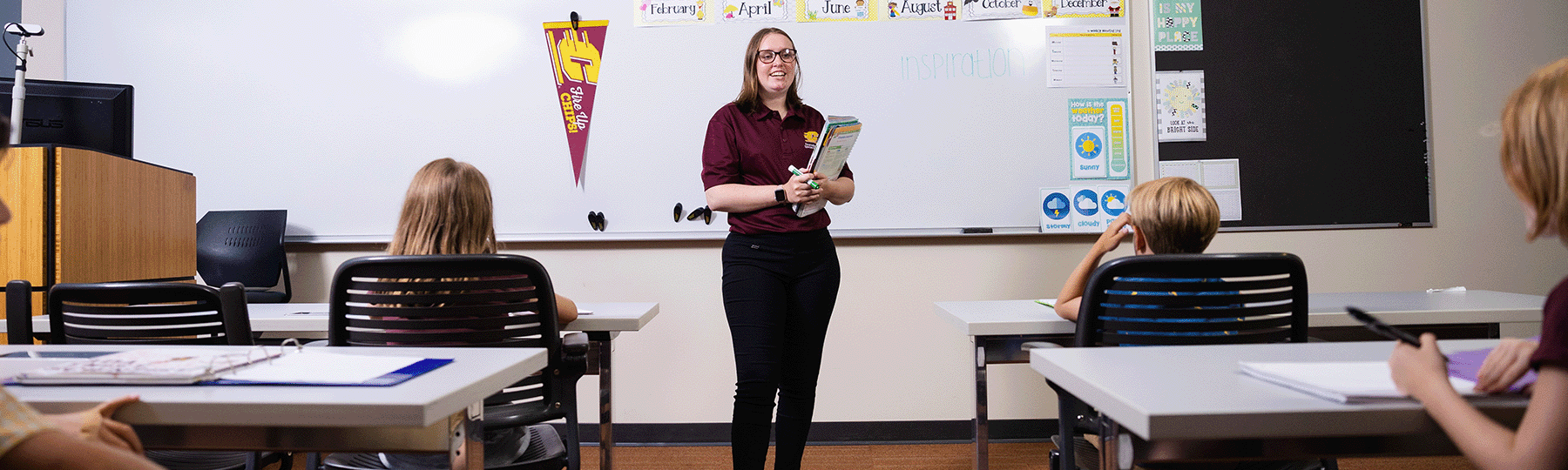 Teacher standing against a whiteboard in front of her students, holding a notebook and smiling while teaching.
