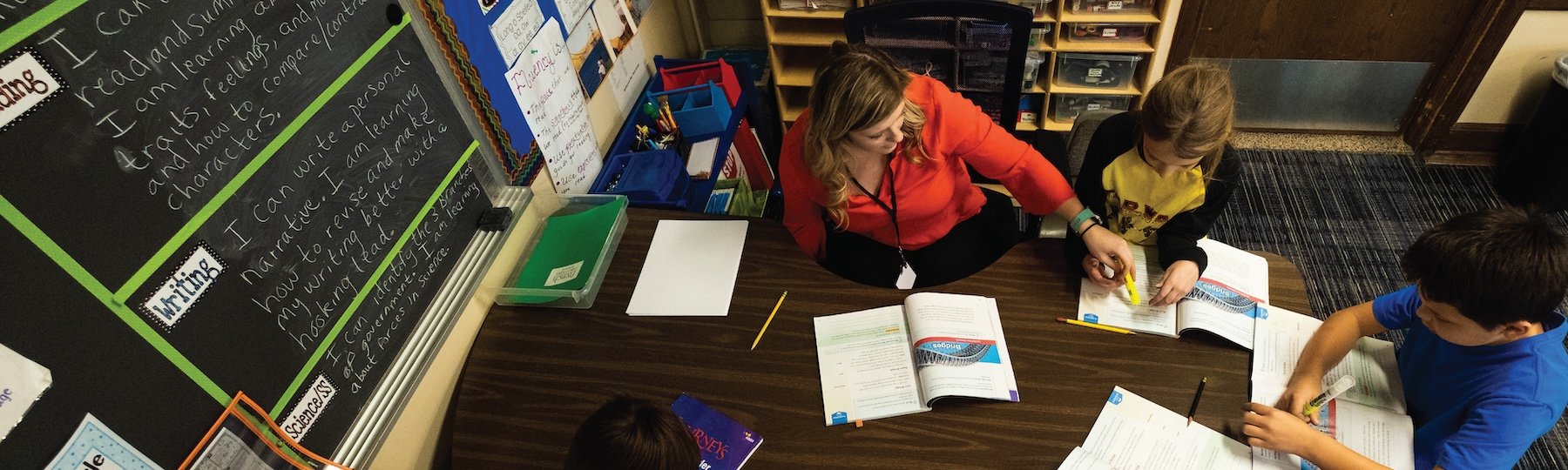 A student teacher wearing an orange shirt, sitting with children at a desk while reading a story book.