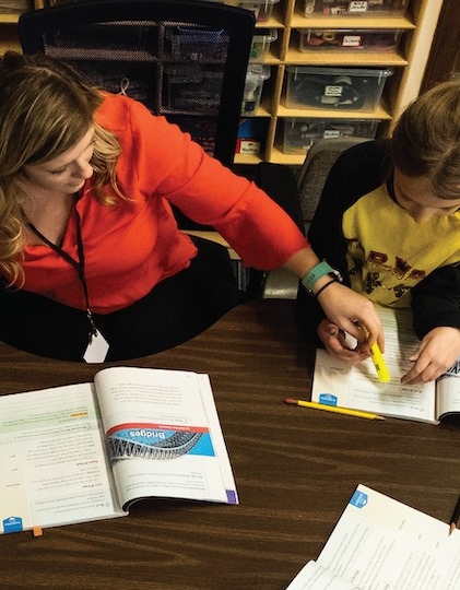 A student teacher wearing an orange shirt, sitting with children at a desk while reading a story book.
