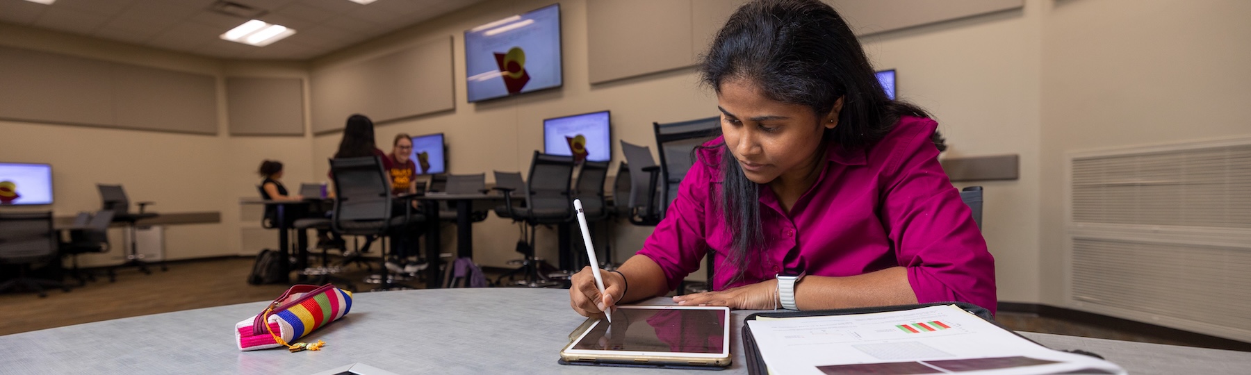 a woman in a pink blouse sits and a table and uses a stylus on a tablet while looking at papers with data charts one them