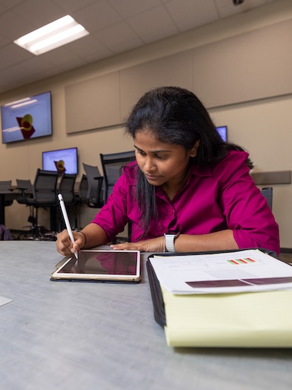 a woman in a pink blouse sits and a table and uses a stylus on a tablet while looking at papers with data charts one them