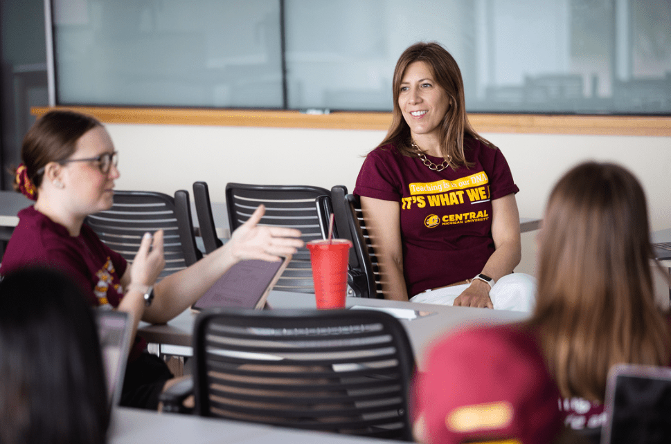 A group of students sitting at a table while talking to an educator. All of them are wearing maroon CMU t-shirts. There is a red cup on the desk as well as paper and an iPad.
