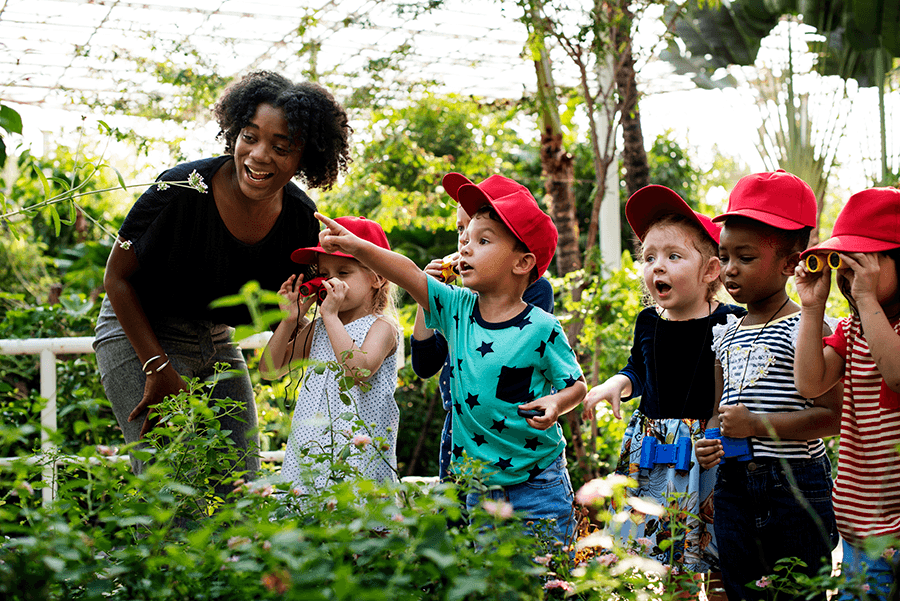 Teacher is talking to a group of young children who are pointing to a plant. The six children are all wearing red hats and are holding binoculars as they look around a greenhouse.