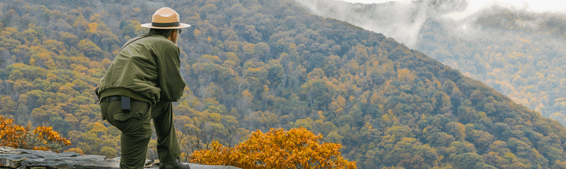 Man wearing a hat looking out into a mountainous range of colorful trees.