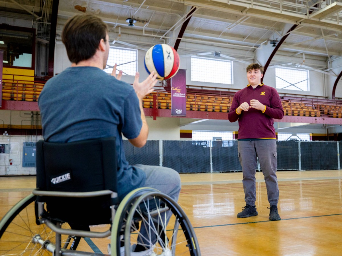 A student in a wheelchair throwing a ball with a student standing up inside a gym area.