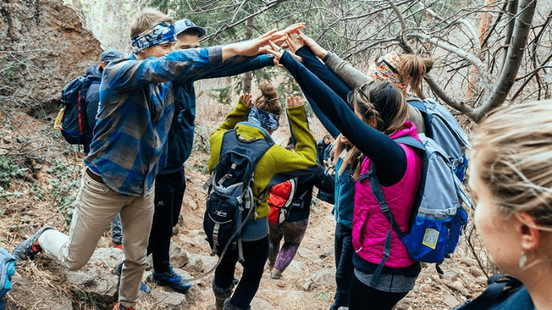 Hikers on a trail standing in a line with their arms forming an archway while another hiker walks underneath them.