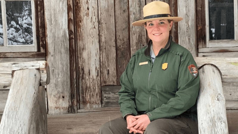 A Park Ranger sitting on steps outside of a cabin wearing a green jacket and tan park ranger hat.