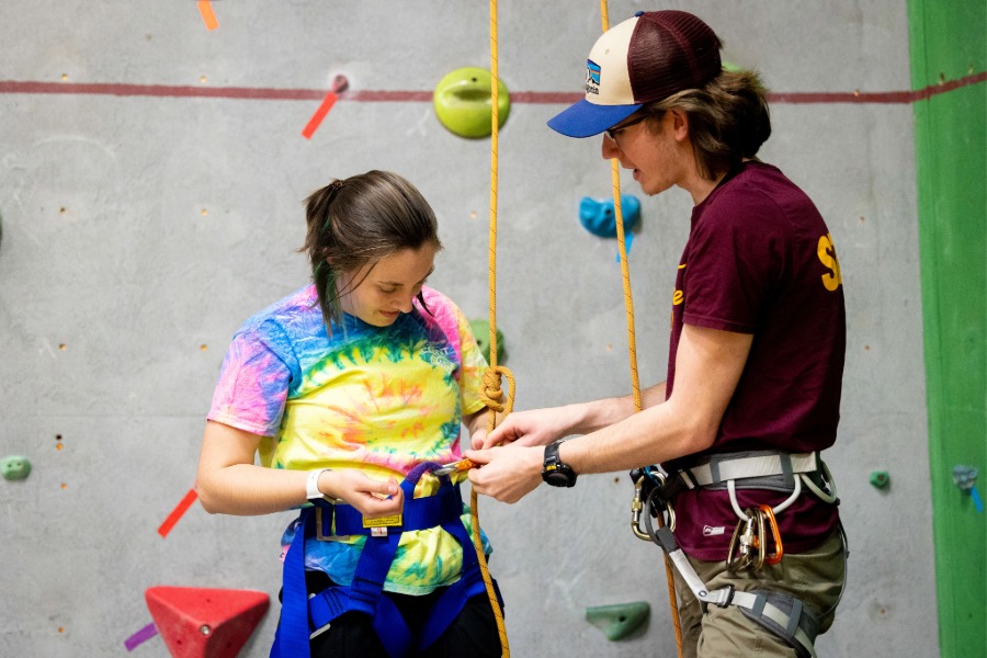 2026-08-Adventure-Seminars-Rock-Wall-01-900x600 A student worker helping another student clip-in to the rope system at the Adventure Seminars rock wall.