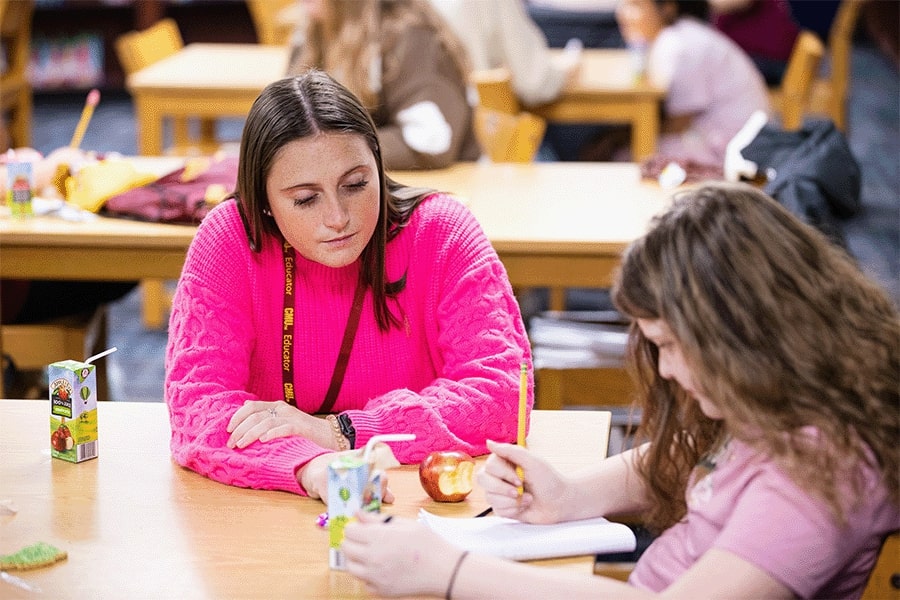 A student teacher sitting at a desk, looking at a young student as she writes in a notebook in a classroom.