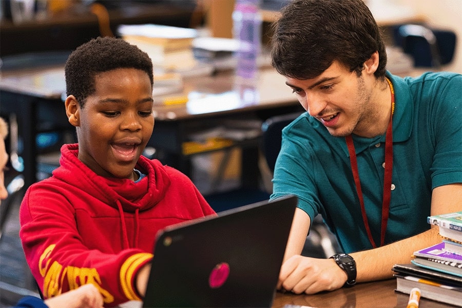 A student teacher sitting at a desk with a young student looking at a computer screen together in a classroom.