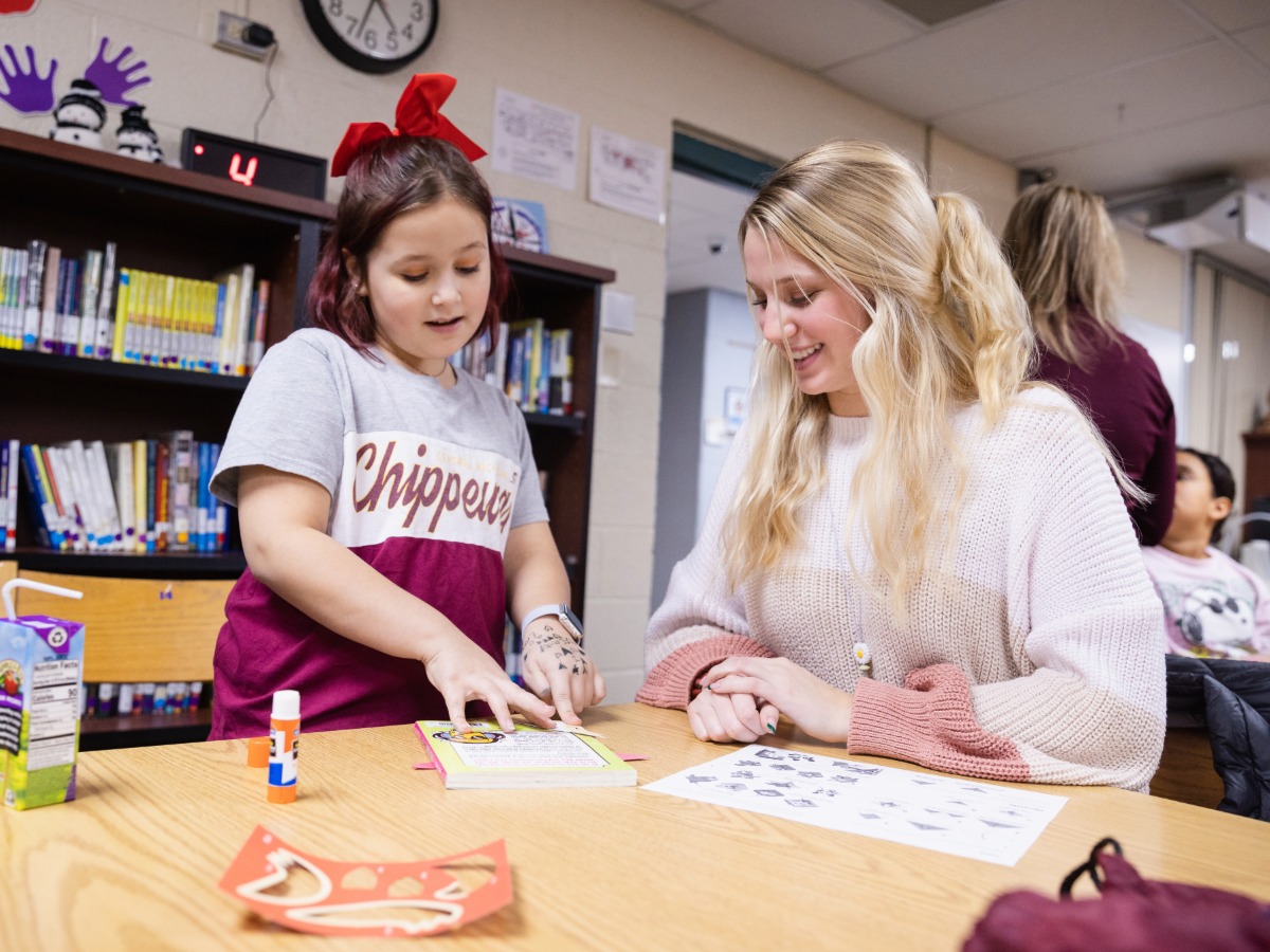 A CMU tutor sitting at a table with a young girl inside of a classroom.