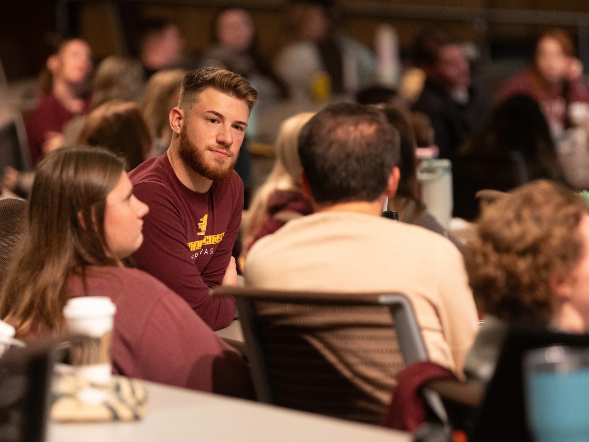 Students sitting at a table with people in the background inside of an auditorium.