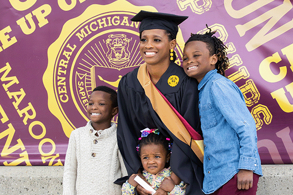 On graduate commencement day an adult female wearing a cap and gown poses with her three proud children.
