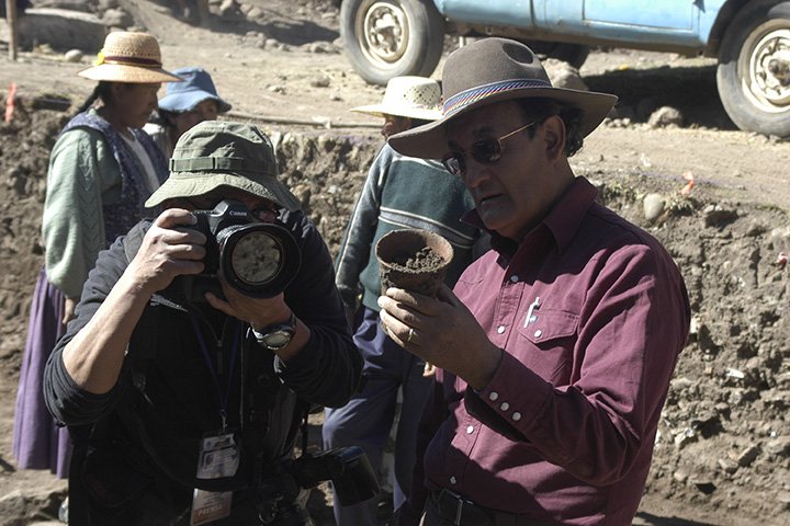 A person wearing a hat holds a large camera while Sergio Chavez, wearing a red shirt and hat, examines an item found at an archaeology research site in the Lake Titicaca Basin of Bolivia.