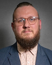 Headshot image of Travis Hill wearing blue jacket over patterned shirt.