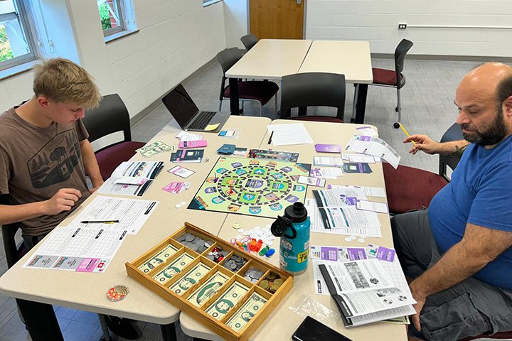 Two individuals sitting at a table with a board game playtest a poverty simulation game.