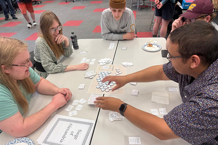 Five students are seated around tables with game boards and pieces in the middle of the tables.