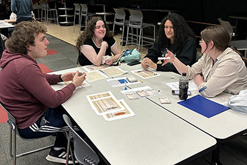 Four students are seated around tables holding playing cards with game boards and cards on the table in front of them.