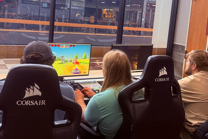 Three students are seated in chairs facing a table with computer monitors and keyboards to play video games.