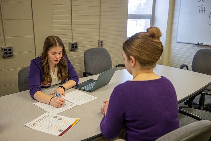 Career peer advisor Kaylynne Costello writes on paper while seated at a table across from a student during office hours at the CLASS Hub.