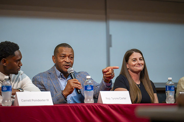 Three individuals sitting at a table with the person in the center holding a microphone.
