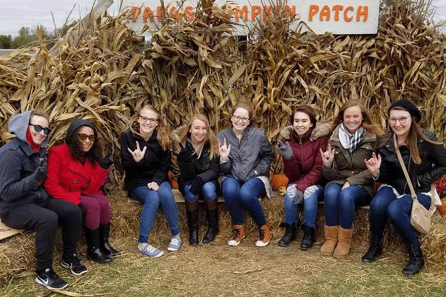 Eight students from the American Sign Language Society at Central Michigan University sit outside at a pumpkin patch on hay with corn stalks around them as they sign.