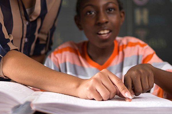 Student reads a Braille book with adult hand guiding child's hand