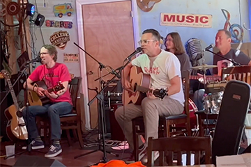 Four band members hold guitars and sing into microphones while seated on a stage.