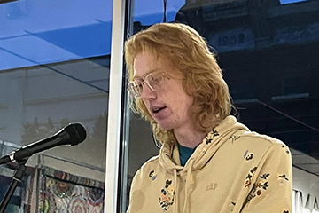 Weston Wise stands at a podium in front of glass windows to read from his poetry at Art Reach of Mid Michigan.
